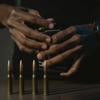 Close-up of dark-skinned hands loading or handling a handgun on a table, with five bullets standing upright and a blue wristwatch visible in the background at right.