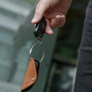 Close-up of a hand gripping a black car key fob with a metal ring and brown leather keychain, against a blurred background.