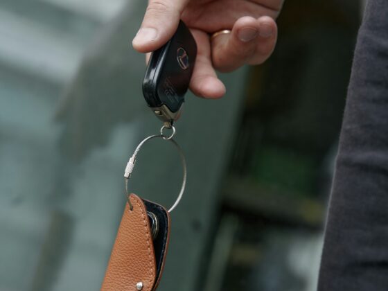 Close-up of a hand gripping a black car key fob with a metal ring and brown leather keychain, against a blurred background.