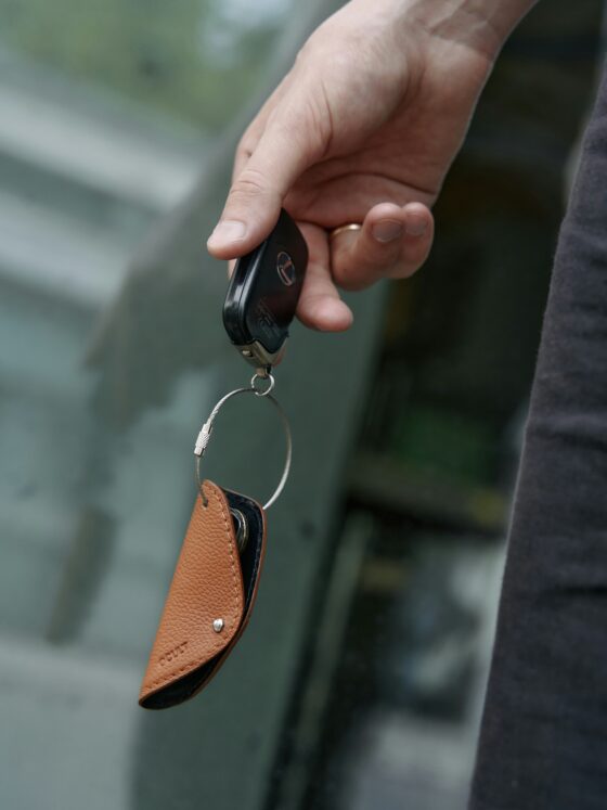 Close-up of a hand gripping a black car key fob with a metal ring and brown leather keychain, against a blurred background.