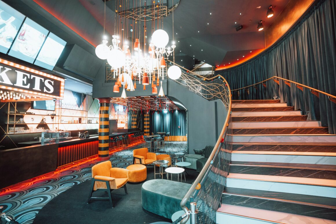 Modern bar and lounge featuring orange seating, a long black counter, and a dramatic chandelier with multiple hanging lights above a patterned floor artfully lit in orange hues.