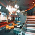 Modern bar and lounge featuring orange seating, a long black counter, and a dramatic chandelier with multiple hanging lights above a patterned floor artfully lit in orange hues.