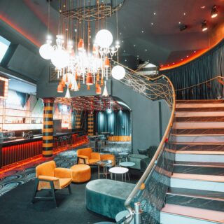 Modern bar and lounge featuring orange seating, a long black counter, and a dramatic chandelier with multiple hanging lights above a patterned floor artfully lit in orange hues.