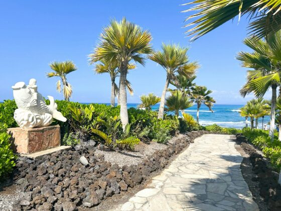 Stone path through a tropical garden with palm trees and a view of the ocean; a white mermaid statue sits on a stone pedestal to the left.