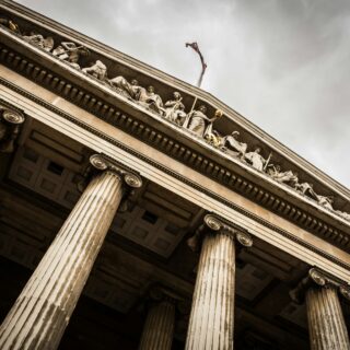 Low-angle view of a neoclassical building with tall fluted columns and a sculpted frieze along the pediment under a cloudy sky