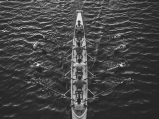 Overhead black-and-white view of a long rowing shell with multiple rowers and oars on calm water.