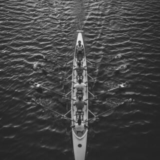 Overhead black-and-white view of a long rowing shell with multiple rowers and oars on calm water.