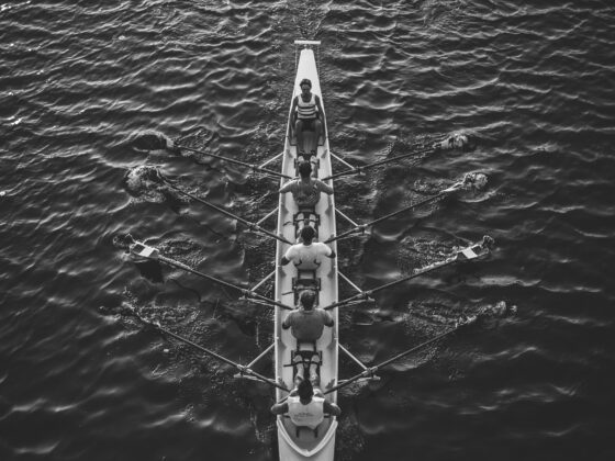 Overhead black-and-white view of a long rowing shell with multiple rowers and oars on calm water.