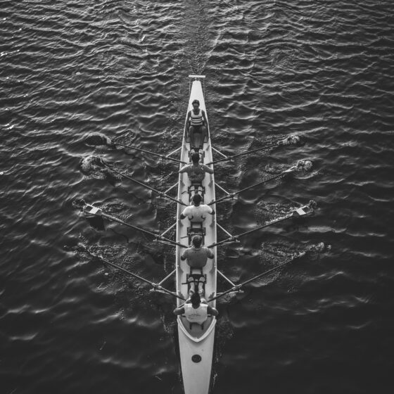 Overhead black-and-white view of a long rowing shell with multiple rowers and oars on calm water.