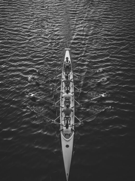 Overhead black-and-white view of a long rowing shell with multiple rowers and oars on calm water.