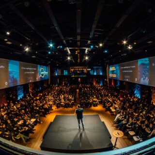Speaker stands on a raised stage addressing a large audience in a dark conference hall with big screens along the sides reading in Portuguese.