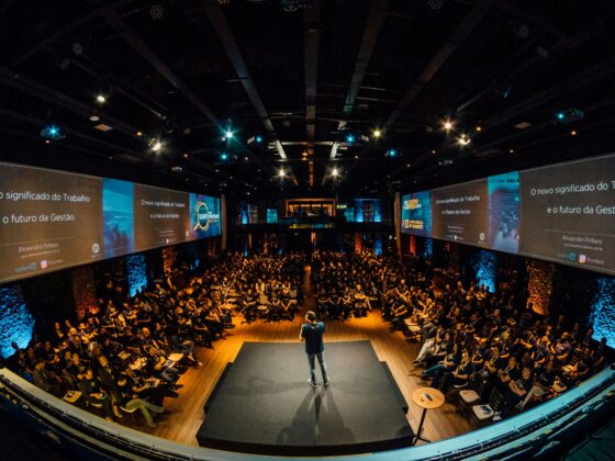 Speaker stands on a raised stage addressing a large audience in a dark conference hall with big screens along the sides reading in Portuguese.