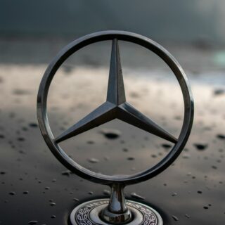 Close-up of a metal Mercedes-Benz emblem on a glossy, water-dotted car hood with a blurred background behind it.