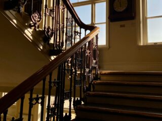 Sunlit interior staircase with a dark wooden handrail and ornate iron balusters, next to a large antique clock on the wall by tall windows.