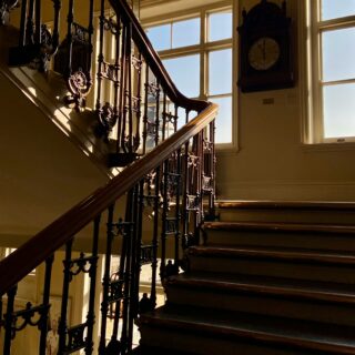 Sunlit interior staircase with a dark wooden handrail and ornate iron balusters, next to a large antique clock on the wall by tall windows.