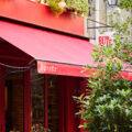 Red storefront café with outdoor seating under a bright red awning; sign reads 'Buvette' with greenery around.