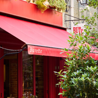 Red storefront café with outdoor seating under a bright red awning; sign reads 'Buvette' with greenery around.