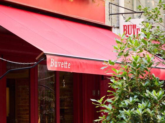Red storefront café with outdoor seating under a bright red awning; sign reads 'Buvette' with greenery around.