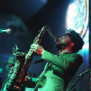 Saxophonist playing a curved saxophone on stage under green and blue concert lighting, microphone nearby.
