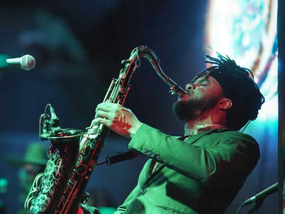 Saxophonist playing a curved saxophone on stage under green and blue concert lighting, microphone nearby.