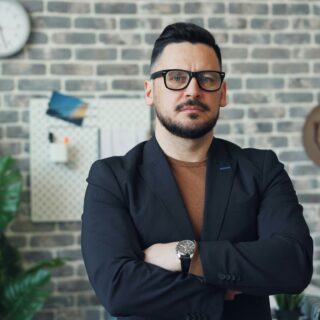 Man in a dark blazer and brown shirt with glasses stands with arms crossed in a brick-walled office.