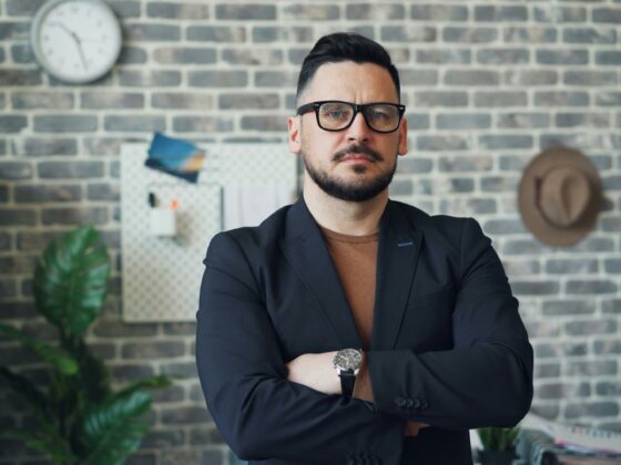 Man in a dark blazer and brown shirt with glasses stands with arms crossed in a brick-walled office.