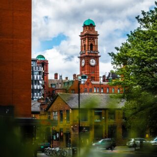 Red brick clock tower with a green copper dome rises above a city street, surrounded by trees and red brick buildings.