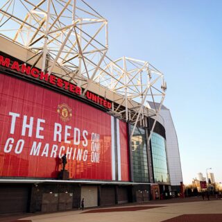Exterior view of Manchester United stadium with a large red banner and white steel framework against a blue sky, capturing the arena's architecture.