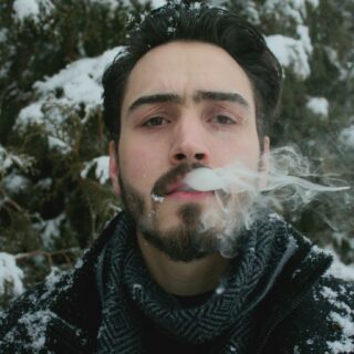 Man with dark hair and beard blowing out a cloud of smoke in a snowy forest setting.
