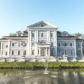 Stately neoclassical building with a central pediment, white façade, and symmetrical windows beside a pond with a fountain.