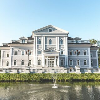 Stately neoclassical building with a central pediment, white façade, and symmetrical windows beside a pond with a fountain.
