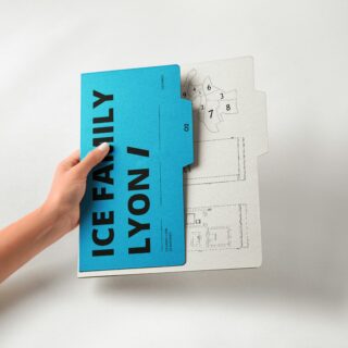 Hand holding a bright blue booklet over blueprint drawings on gray paper. It emphasizes the booklet atop architectural plans due to the visible hand and overlapping sheets.