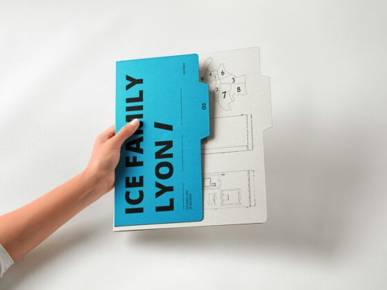 Hand holding a bright blue booklet over blueprint drawings on gray paper. It emphasizes the booklet atop architectural plans due to the visible hand and overlapping sheets.