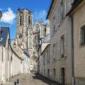 Narrow cobblestone street toward a Gothic cathedral with intricate stonework and rose window, beige buildings with white shutters on both sides, blue sky above