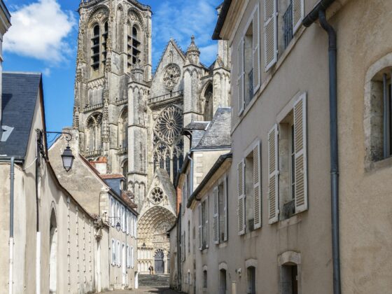 Narrow cobblestone street toward a Gothic cathedral with intricate stonework and rose window, beige buildings with white shutters on both sides, blue sky above