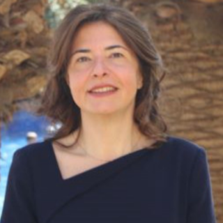 Portrait of a woman with shoulder-length brown hair, wearing a dark blue top, standing outdoors with palm trees and a pool in the background.