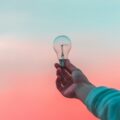 A hand holding a clear light bulb up against a pastel sunset sky with pink and teal tones.