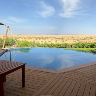 Wooden deck surrounds a swimming pool with a shade sail and desert landscape in the distance under a clear blue sky