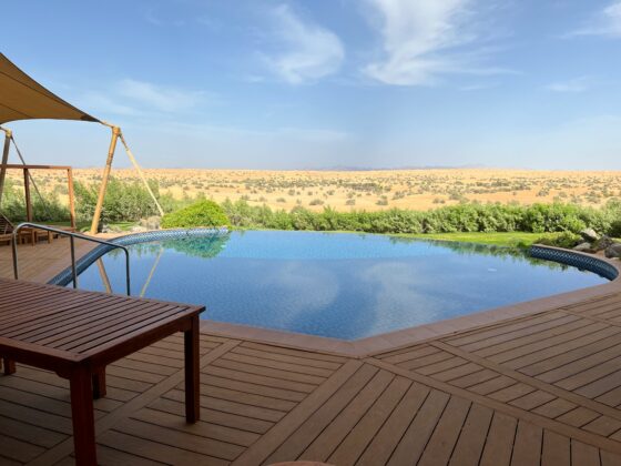Wooden deck surrounds a swimming pool with a shade sail and desert landscape in the distance under a clear blue sky