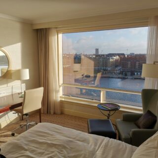 Hotel room with a large window view of a river and city skyline; round wall mirror, desk, and cream furniture in the foreground.