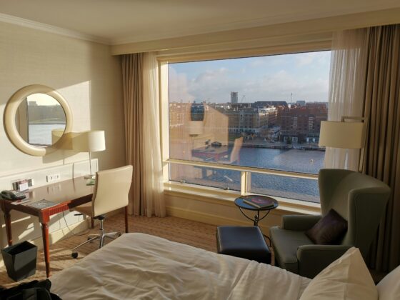 Hotel room with a large window view of a river and city skyline; round wall mirror, desk, and cream furniture in the foreground.
