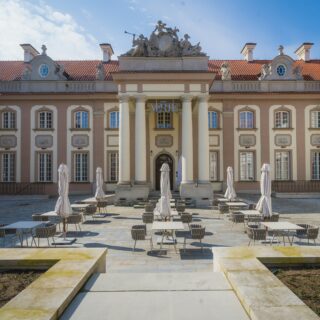 Courtyard of a historic hotel with a central portico and tall columns, rows of tables and chairs with closed umbrellas, beige building façades and a red-tiled roof.