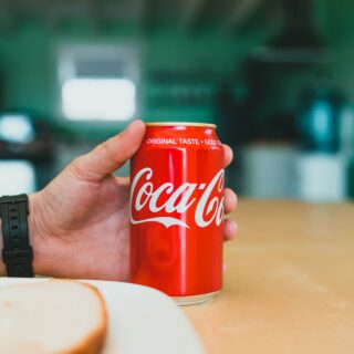 Close-up of a hand holding a red Coca-Cola can, foreground in focus and a blurred green background behind it.