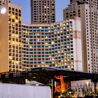 Evening view of a large beige hotel complex with lit windows and balconies, pool area with umbrellas in the foreground.