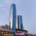 Tall curved glass skyscraper reflecting the blue sky, with lower modern buildings nearby.
