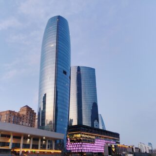 Tall curved glass skyscraper reflecting the blue sky, with lower modern buildings nearby.