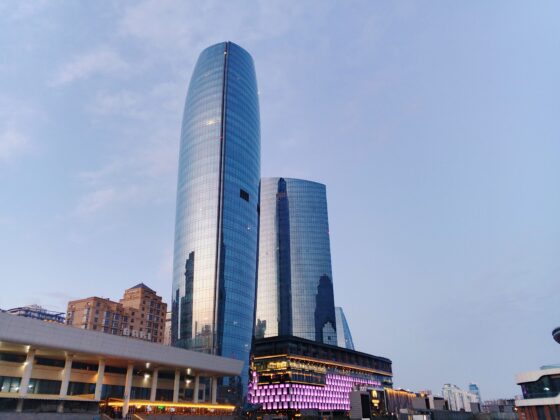 Tall curved glass skyscraper reflecting the blue sky, with lower modern buildings nearby.