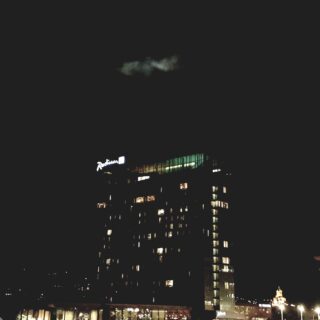 Nighttime cityscape with illuminated Radisson sign on a curved glass building and a bright, geometric-lit tower to the right against a dark sky.