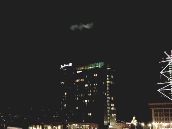 Nighttime cityscape with illuminated Radisson sign on a curved glass building and a bright, geometric-lit tower to the right against a dark sky.