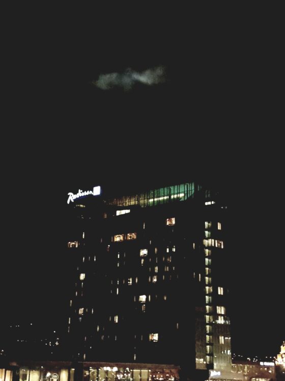 Nighttime cityscape with illuminated Radisson sign on a curved glass building and a bright, geometric-lit tower to the right against a dark sky.
